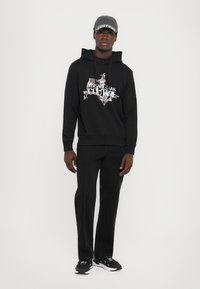 Um hoodie preto com uma estampa gráfica em prateado, combinado com calças pretas com costuras contrastantes e ténis pretos. Completa o visual com um chapéu cinza.