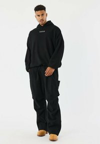 Sudadera negra con capucha y un parche de logo, combinada con pantalones cargo negros con bolsillos laterales y calzado plano color marrón claro.