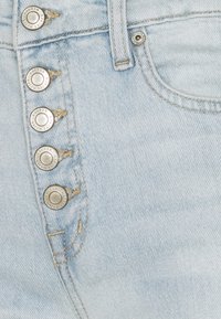 Ljusblå denimjeans har en hög midja, fem blanka metallknappar och synliga sömmar längs midjebandet.