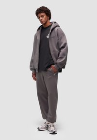 Felpa grigia oversize con zip frontale, t-shirt nera sotto, e pantaloni del pigiama grigi coordinati. Le sneakers presentano una finitura argentata.