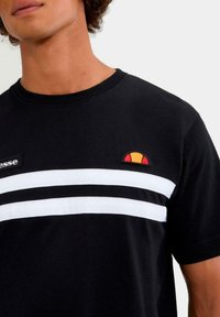 Maglietta nera a righe orizzontali bianche, con un piccolo logo colorato e una toppa con il nome del marchio. Materiale in cotone con texture liscia.