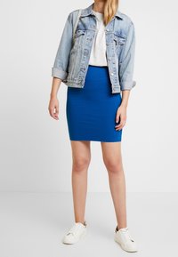 Jupe bleu moulante associée à une veste en denim clair, un t-shirt gris et des baskets blanches. La jupe est en tissu doux et extensible.