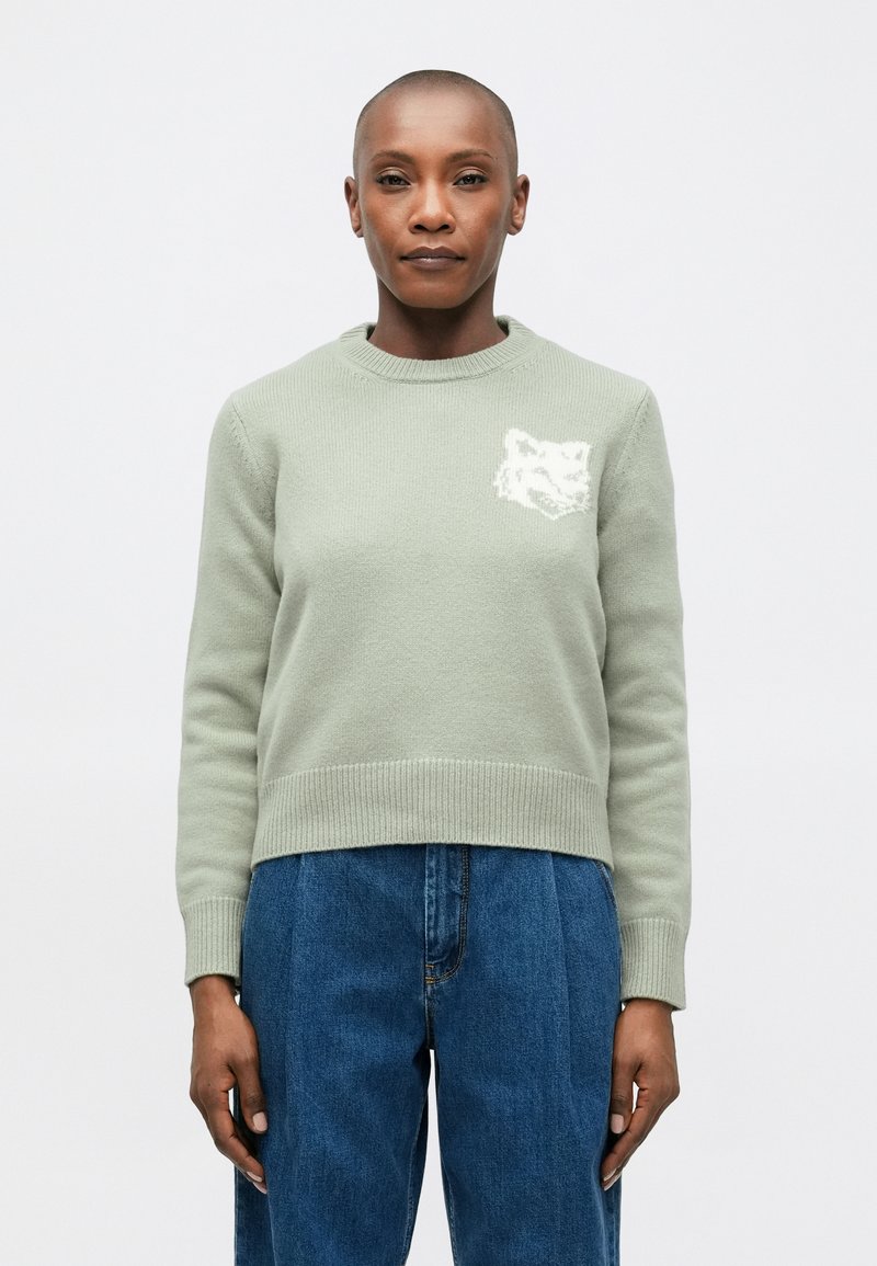 Suéter de punto verde claro con cuello redondo y puños acanalados, con un gráfico de un zorro blanco en el lado izquierdo del pecho. Combinado con pantalones azul de tiro alto.