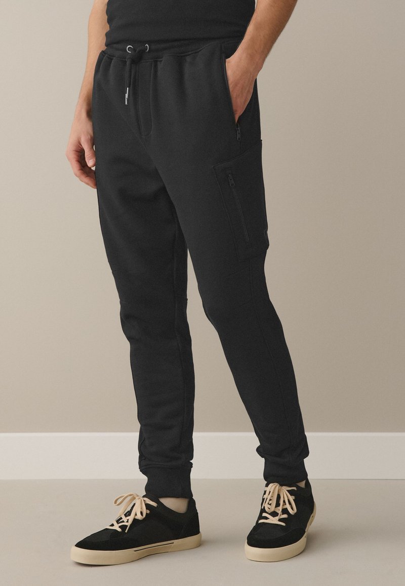 Uomo con pantaloni jogger neri con tasche laterali con zip e scarpe casual nere con suola e lacci beige, in piedi contro una parete beige.
