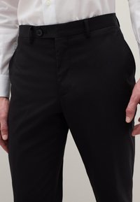 Zwarte pantalon met een gladde textuur, taps toelopende pasvorm, twee voorzakken en een enkele knoopsluiting aan de tailleband, gecombineerd met een wit overhemd.