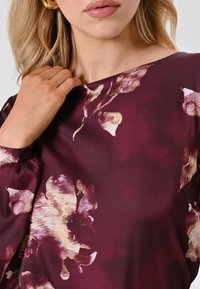 Bluse mit floralem Muster in tiefem Burgunderrot und hellen Akzenten. Besitzt einen Bootsausschnitt und sanft geraffte Ärmel. Glatte Stoffoberfläche.