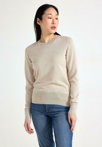Maglione beige lavorato a maglia con maniche lunghe e collo tondo, caratterizzato da un orlo e polsini a costine. Indossato con jeans blu, evidenziando uno stile casual.