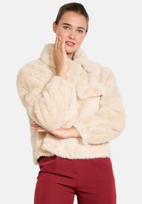 Jakna iz umetnega krzna v kremni barvi s kragnim izrezom, skrajšanim dizajnom in teksturiranimi poudarki, v kombinaciji z burgundskimi hlačami.
