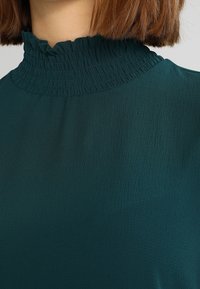 Blusa teal con un colletto alto, arricciato e smockato, realizzata in un tessuto liscio. Presenta una leggera lucentezza e una vestibilità rilassata.