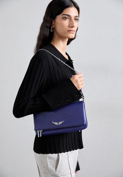 Purple leather crossbody bag with a silver wing-shaped accent, featuring a zipper closure and a chain strap, worn with a black ribbed shirt.