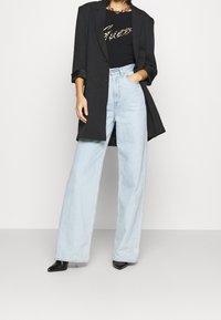 Svart blazer över en svart topp med ett guldmönster, kombinerat med ljusblå vida jeans och svarta spetsiga klackar. Släta tyger och strukturerad design.