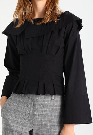 Kobieta ubrana w czarną bluzkę z długim rękawem, szerokim kołnierzem i plisowanym pasem, zestawioną z szarymi spodniami w kratę o wysokim stanie.