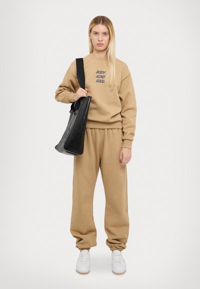 Beige sweatshirt with "BODY MIND SOUL" text, matching sweatpants, and white sneakers. Carrying a black bag with a smooth texture.
