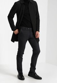 Cappotto nero con colletto, indossato sopra un dolcevita nero aderente. Pantaloni grigi slim-fit con un motivo sottile, abbinati a sneaker nere.