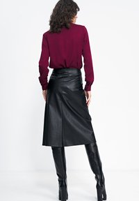 Czarna skórzana spódnica o wysokim staniku, długości midi i gładkiej fakturze, zestawiona z bordową bluzką z długim rękawem. Kasztanowe włosy, widok z tyłu.