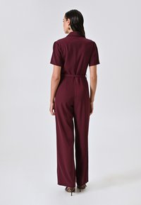 Tutina bordeaux con maniche corte, vita cintrata, gambe larghe e texture morbida. Abbinata a tacchi, vista da dietro.