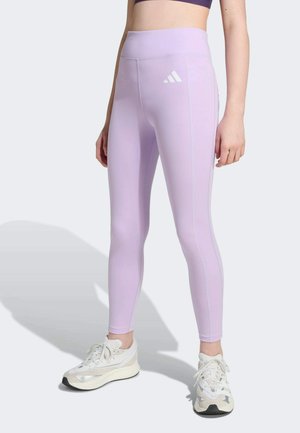 Leggings lilas clairs, taille haute, fabriqués en tissu extensible. Présentent un logo blanc sur la cuisse gauche et des accents sur les coutures latérales. Portés avec des baskets blanches.
