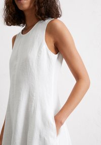 White sleeveless dress with a textured fabric, featuring side pockets and a rounded neckline, emphasizing a clean and minimalist design.