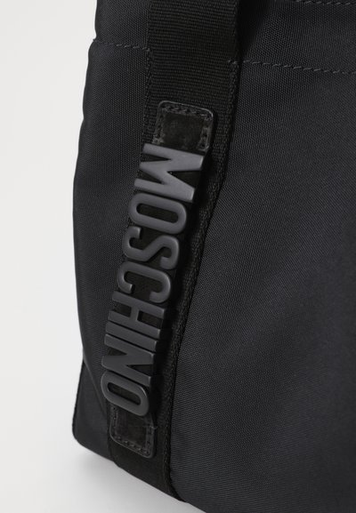 Sac en nylon noir présentant une surface texturée, avec un logo Moschino en plastique surélevé sur le côté et une bandoulière en tissu robuste.