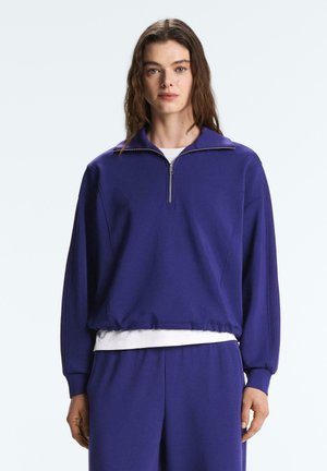 Giovane donna con capelli castani lunghi che indossa un pullover viola con cerniera e pantaloni coordinati, in piedi davanti a uno sfondo bianco.