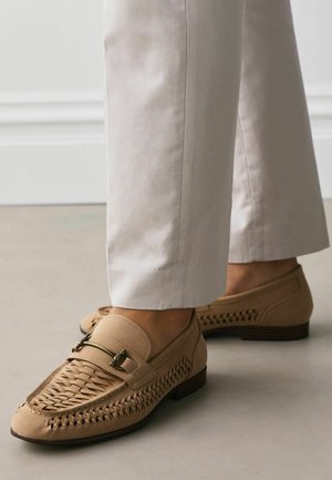 Pieds portant des mocassins en cuir tressé beige avec détail boucle métallique et pantalons droits de couleur claire sur un sol lisse.