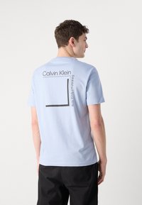 Lysblå bomulds t-shirt med et sort grafisk tryk på ryggen, der har teksten "Calvin Klein" og "Established Since 1978". Korte ærmer.
