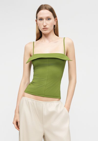 Νεαρή γυναίκα με ίσια μαλλιά φοράει ένα πράσινο στενό τοπ χωρίς μανίκια με λεπτές τιράντες και μπεζ παντελόνι ψηλόμεσο, στέκεται με το ένα χέρι στην τσέπη.