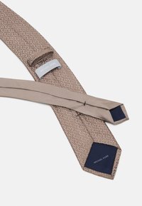 Beige slips med et subtilt geometrisk mønster, glat tekstur og marineblå accenter. Har et mærke med "MICHAEL KORS" på bagsiden.