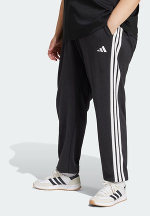 Osoba w czarnych spodniach dresowych Adidas z białymi bocznymi paskami i białych butach sportowych Adidas z czarnymi paskami, stojąca z jedną ręką w kieszeni.