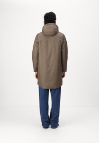 Long, taupe, waterproof jacket with a hood, featuring a straight cut and side slits, worn over blue, wide-leg jeans. Smooth texture, minimalist design.
