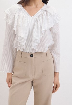 Vrouw in een witte blouse met pinstripes en gelaagde ruches, gecombineerd met beige hoge taille broeken met voorzakken en een zwarte knoop.