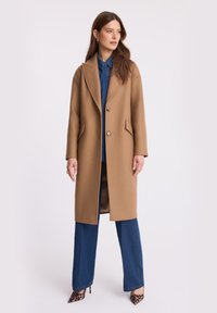 Kobieta ubrana w długi płaszcz w kolorze camel, niebieską koszulę dżinsową oraz spodnie, a także wysokie obcasy w leopardzie wzory, stojąca na jednolitym tle.