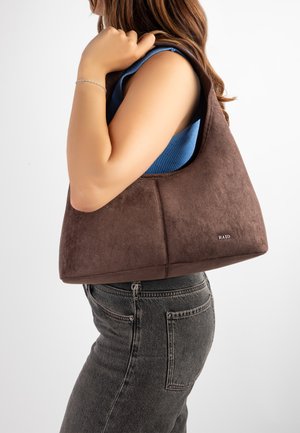 Femme portant un haut bleu sans manches et un jean noir, portant un grand sac à main en daim marron sur l'épaule sur fond blanc.