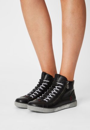 Jambes portant des baskets montantes noires avec des lacets blancs, une fermeture éclair latérale et des semelles en caoutchouc grises texturées sur un fond uni.