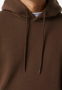 Hnědá mikina s kapucí s hladkým povrchem, s táhly se stříbrnými konci a žebrovaným výstřihem. Jednoduchý, ležérní design.