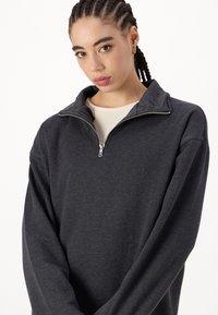 Siva zip-up dukserica z visokim ovratnikom in dolgimi, širokimi rokavi. Izdelana iz mehkega materiala, z rahlo teksturo in kovinskim zadrgo.