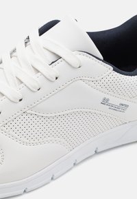 White leather sneaker with perforated design, lace-up closure, and navy blue accents on the tongue. Features a textured sole for grip.
