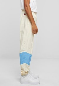 Pantaloni de trening bej cu un model în zigzag albastru pe picioare. Realizați dintr-un material ușor, cu un bandă elastică la talie. Se poartă adidași albi.