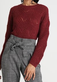 Czerwony sweter na drutach z falistym wzorem, zestawiony z wysokim stanem szarymi spodniami w kratę z wiązanym paskiem.