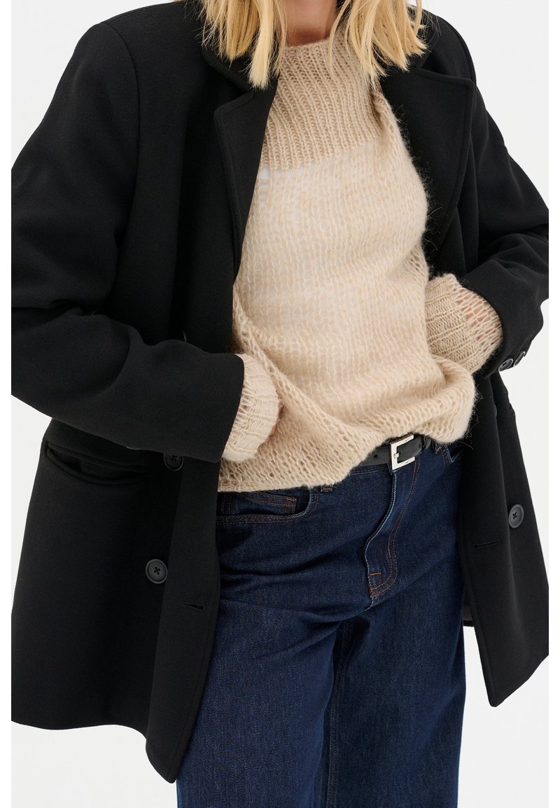Blazer nero realizzato in tessuto testurizzato, caratterizzato da grandi bottoni e un taglio sartoriale, indossato sopra un maglione di pelliccia beige chiaro e jeans in denim scuro.