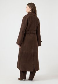 Brązowy płaszcz ze skóry zamszowej z paskiem w talii, długimi rękawami i długością do połowy łydki. Posiada rozcięcie z tyłu dla łatwości ruchu.