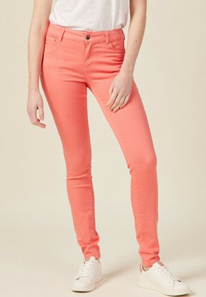 Schmal geschnittene korallenfarbene Hose, kombiniert mit weißem Oberteil und weißen Sneakers, von der Taille bis zu den Füßen vor einfarbigem Hintergrund gezeigt.