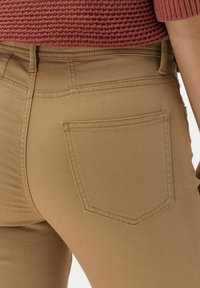 Pantalones beige con bolsillo trasero y trabillas, llevados con un suéter de punto texturizado color óxido, toma en primer plano de la parte baja de la espalda y la cintura.