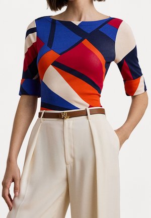 Frau trägt ein farbenfrohes Oberteil mit geometrischem Muster in Blau, Rot, Orange und Beige, mit ellbogenlangen Ärmeln und eine beige hoch taillierte Hose mit braunem Gürtel.