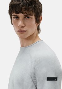 Lichtgrijs langärmelige shirt van textuurstof met geribbelde schouderaccenten; heeft een zwart label op de linkermouw.