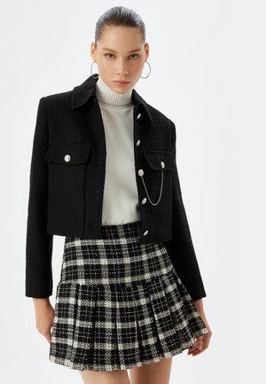 Zwarte tweed cropped jasje met knoopsluiting en voorzakken, gecombineerd met een witte geribbelde coltrui en een geplooide zwart-wit geruite rok.