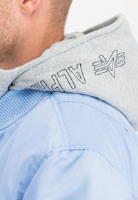 Close-up van een persoon die een lichtblauw jack draagt met een grijze capuchon waarop zwarte "ALPHA" tekst en logo geborduurd zijn.