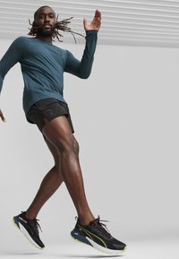 Sportiniai batai su juodu tekstūruotu viršumi; atspindinčios detalės, žalias padas. Modelis vilki mėlyną ilgomis rankovėmis marškinėlį ir juodas šortus, viduryje bėgimo.