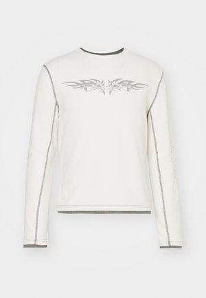 Biała koszula z długim rękawem o prążkowanej strukturze, z szarymi wzorami plemiennymi z przodu i szarym przeszyciem wzdłuż szwów i mankietów.