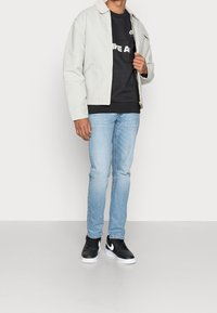 Ljust grå zip-up-jacka med en beige krage, mörk sweatshirt med logotyp, blå jeans och svarta sneakers med vita accent. 